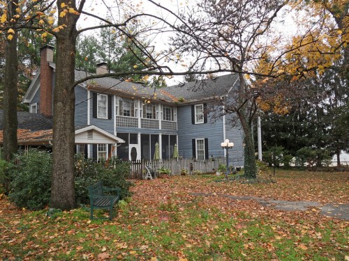 A large blue house surrounded by autumn leaves and trees, featuring a porch and a bench in the yard.