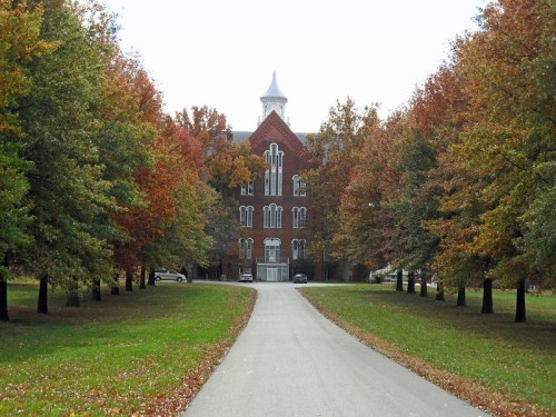 A tree-lined path leads to a historic brick building surrounded by autumn foliage.