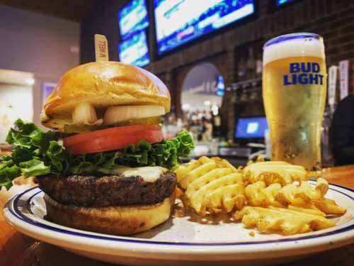 A juicy burger with lettuce, tomato, and onion, served with waffle fries and a pint of Bud Light in a bar setting.