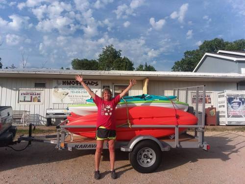 A person in a red shirt stands with arms raised beside a trailer loaded with colorful kayaks in front of a building.