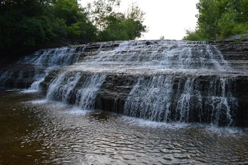 A serene waterfall cascading over rocky steps into a calm pool, surrounded by lush greenery.