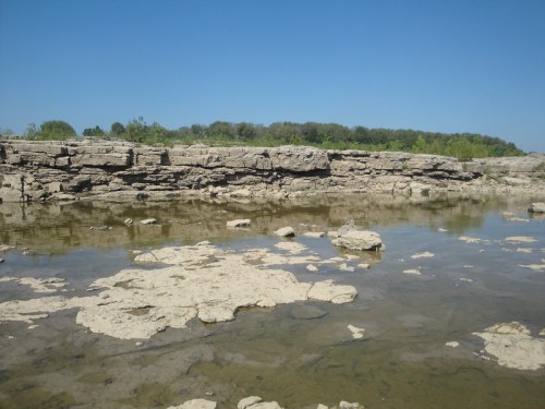 Rocky landscape with shallow water pools under a clear blue sky and green trees in the background.