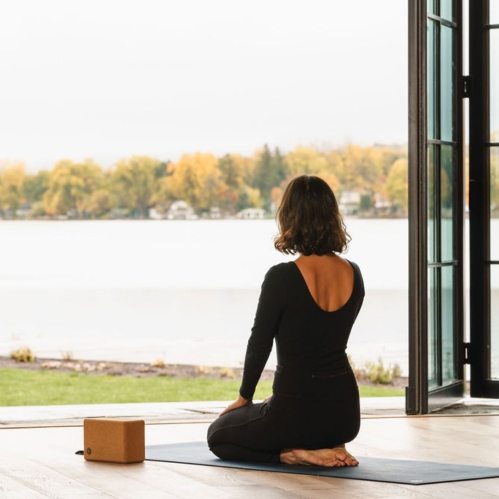 A woman practices yoga by a lake, sitting on a mat with a block beside her, surrounded by autumn scenery.