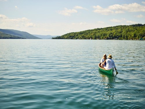 A couple paddles a small canoe on a calm lake surrounded by green hills under a clear blue sky.