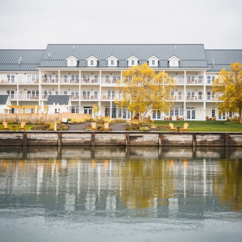 A waterfront hotel with a gray roof, surrounded by greenery and autumn trees, reflecting in the calm water.