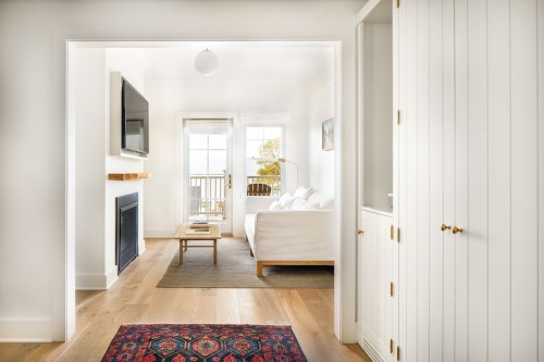 Bright, modern living room with a white sofa, TV, and balcony access, featuring light wood floors and a colorful rug.