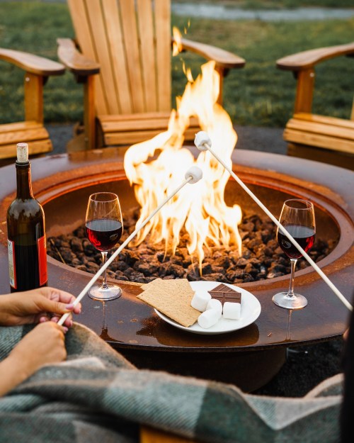 A cozy fire pit scene with two glasses of red wine, s'mores ingredients, and someone roasting marshmallows.