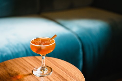A cocktail in a coupe glass with a cinnamon stick, resting on a wooden table beside a soft, blue couch.
