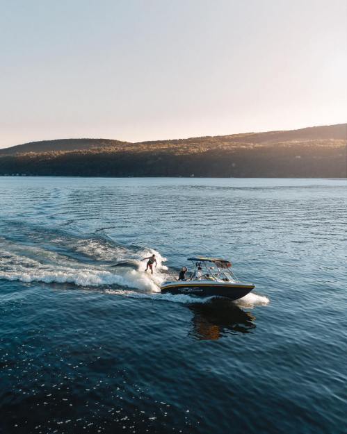 A boat with two people surfing behind it on a calm lake, surrounded by hills at sunset.