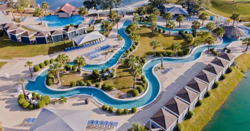 Aerial view of a resort with a winding lazy river, palm trees, and poolside loungers near a tranquil water body.