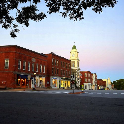 A quiet street scene featuring historic brick buildings and a clock tower at dusk, with a clear sky in the background.