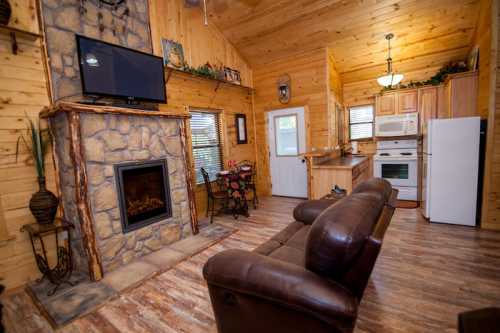 Cozy cabin interior with wooden walls, a stone fireplace, a TV, and a kitchen area featuring a dining table.