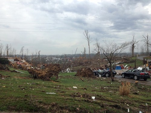 Devastated landscape after a tornado, with downed trees, debris, and damaged vehicles under a cloudy sky.