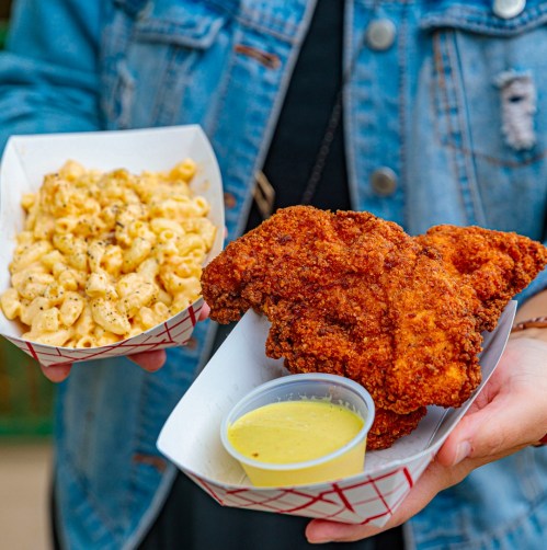 A person holds two food trays: one with crispy fried chicken and a side of mac and cheese, the other with dipping sauce.