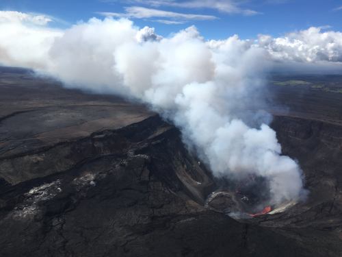 Aerial view of a volcano emitting smoke and lava, surrounded by a barren landscape and blue sky with clouds.