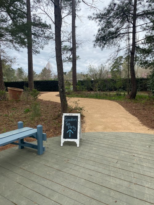 A wooden sign points to a garden tea party, with a winding path and trees in the background.