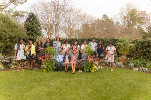 A group of twelve people poses together in a lush garden, surrounded by greenery and colorful plants.