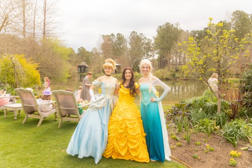 Three women in princess costumes stand together by a pond, surrounded by greenery and children playing in the background.