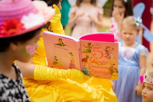 A person in a yellow dress reads "5 Minute Princess Stories" to a group of children in colorful costumes.