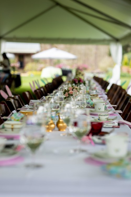 A beautifully set outdoor dining table under a tent, adorned with flowers and elegant tableware.