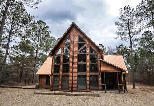 A modern wooden cabin with large windows, surrounded by trees and a gravel pathway under a cloudy sky.