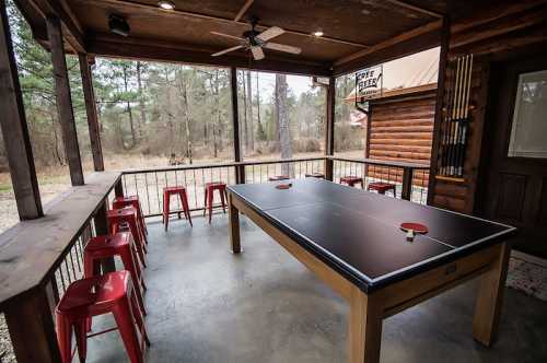 A covered outdoor space featuring a ping pong table and red stools, surrounded by trees and nature.