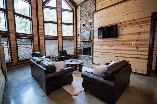 Cozy living room with large windows, stone fireplace, leather sofas, and wooden walls, creating a warm atmosphere.