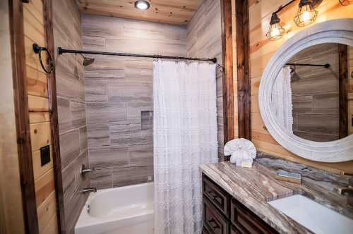 A cozy bathroom featuring a shower with a curtain, wooden accents, and a round mirror above a stone countertop.