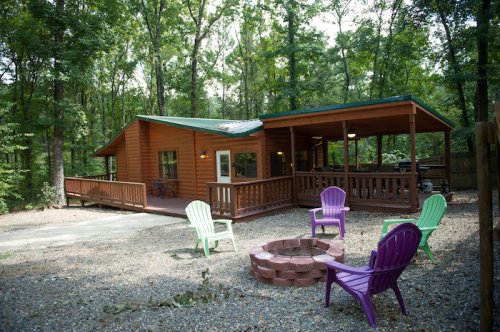 A cozy wooden cabin surrounded by trees, featuring a porch and a fire pit with colorful chairs in the foreground.