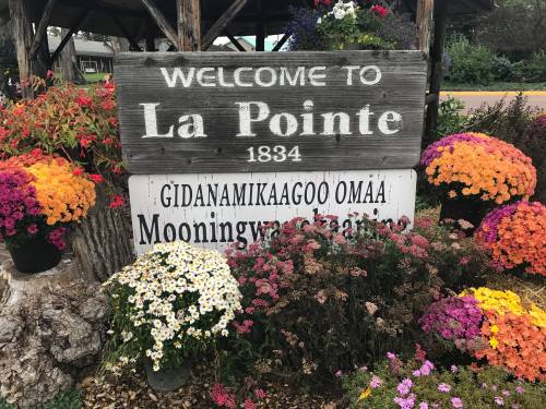 A wooden sign reading "Welcome to La Pointe 1834" surrounded by colorful flowers and plants.