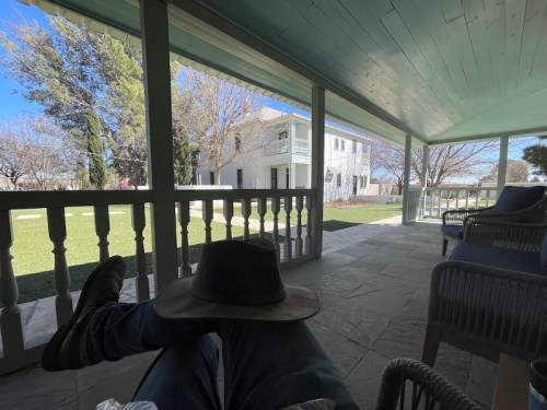 A person relaxes on a porch, wearing a hat, with a view of a grassy yard and a white house in the background.