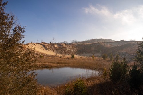 A serene landscape featuring sand dunes, a calm pond, and sparse vegetation under a clear blue sky.