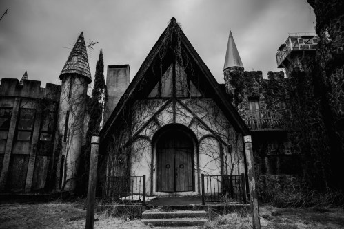 A dark, abandoned building with a steep roof, surrounded by overgrown vines and stone towers under a cloudy sky.
