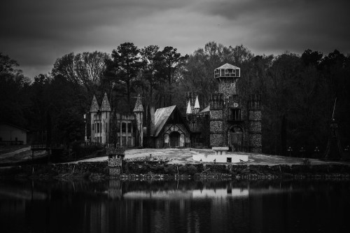 A dark, eerie castle with towers and a reflective lake, surrounded by trees under a cloudy sky.