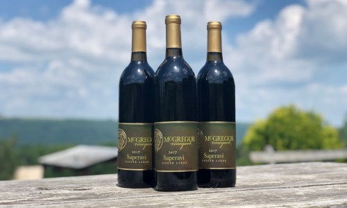 Three bottles of 2017 McGregor Vineyard Saperavi wine on a wooden table with a scenic background.