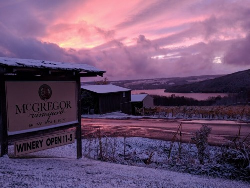A sign for McGregor Vineyard & Winery with a stunning sunset over a snowy landscape and a lake in the background.