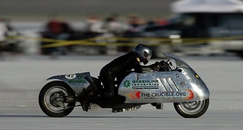 A rider on a sleek, silver motorcycle speeds across a flat, sandy surface at a racing event.