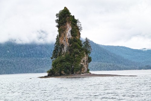 A tall, rocky island covered in greenery rises from calm waters, surrounded by misty mountains and cloudy skies.