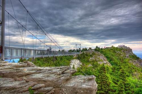 A suspension bridge spans a rocky landscape, surrounded by lush greenery and under a dramatic, cloudy sky.