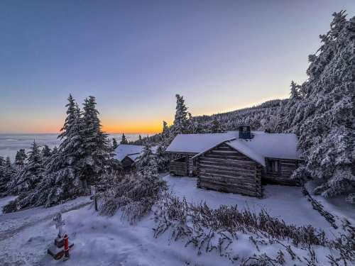 Snow-covered cabins surrounded by frosty trees at sunset, with a serene landscape in the background.