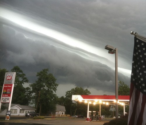 Dramatic storm clouds loom over a gas station, with an American flag in the foreground and trees in the background.