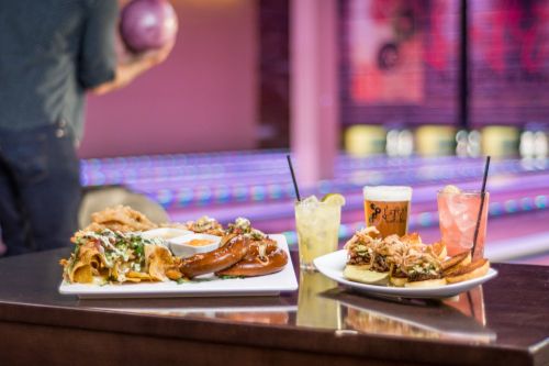 A table with two plates of food and drinks, set against a bowling alley backdrop. One plate features nachos and sausage.