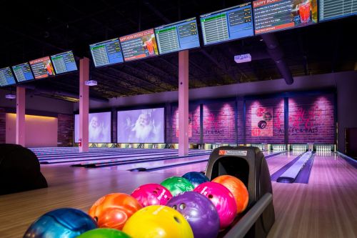 A bowling alley with colorful bowling balls in the foreground and screens displaying scores in the background.