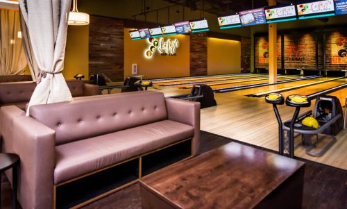A cozy bowling alley lounge with a brown sofa, wooden tables, and bowling lanes in the background.