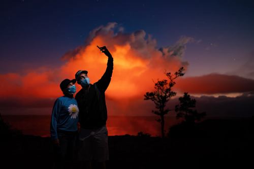 A couple wearing masks poses for a selfie against a vibrant sunset and volcanic cloud backdrop.