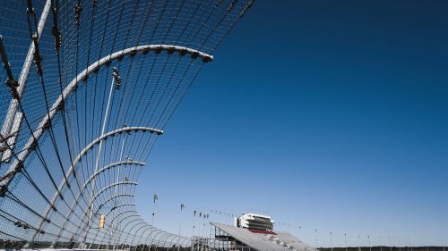 A racetrack with a tall safety fence and grandstands under a clear blue sky.