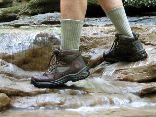 A person wades through a shallow stream, wearing brown hiking boots and light green socks.