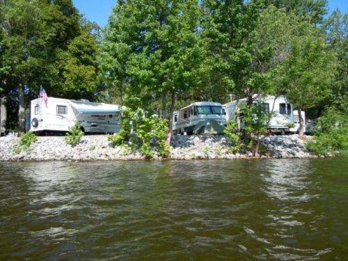 Three RVs parked by a rocky shoreline, surrounded by green trees, with a clear blue sky above and water in the foreground.