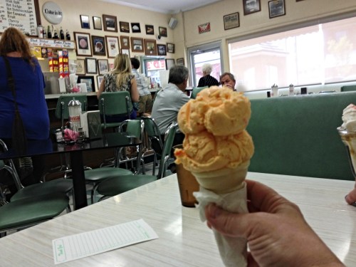 A hand holds a cone with three scoops of orange ice cream in a retro ice cream shop filled with patrons.
