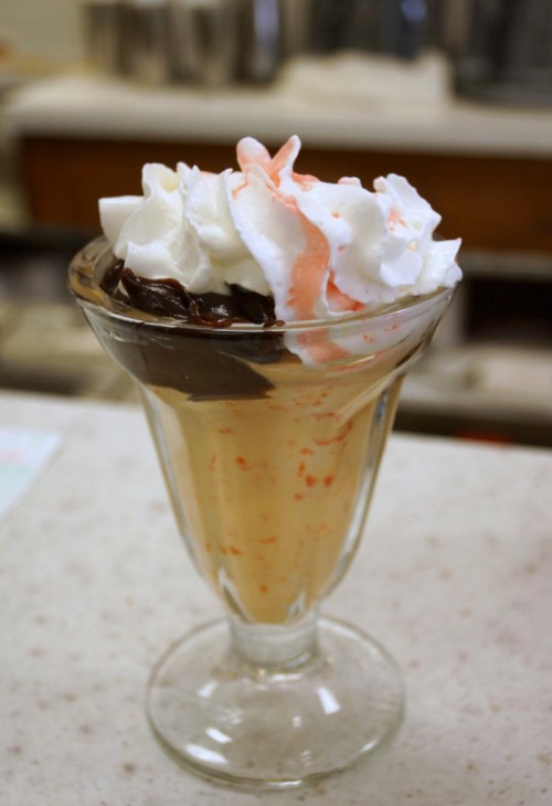 A glass dessert featuring layers of vanilla ice cream, chocolate sauce, and whipped cream topped with a drizzle of strawberry.